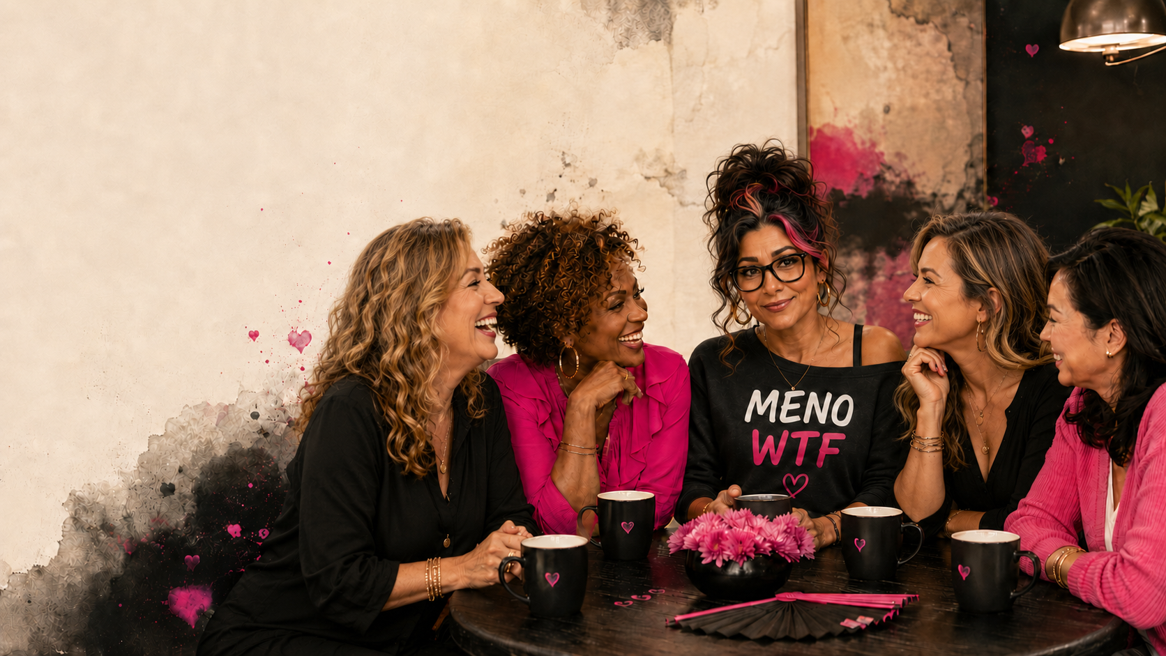 Five women laughing together around a table with mugs, wearing MENO WTF apparel